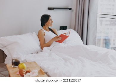 Side View Profile Cheery Young Woman In White Tank Top Sit In Bed Reading Book Looking Aside Rest Relax Spend Time In Bedroom Lounge Home In Own Room House Wake Up Dream Be Lost In Reverie Good Day