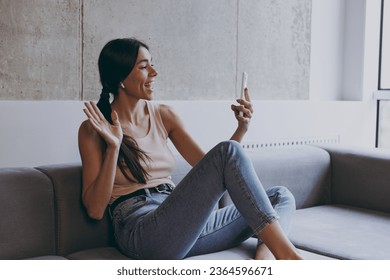 Side view profile fun young latin woman 20s in casual clothes sit on sofa in earphones get video call using mobile cell phone talk greet with hand spend time in living room home in own house good day