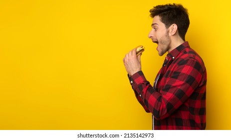 Side View Profile Portrait Of Funny Hungry Guy Holding Biting Burger Eating Junk Food Posing With Open Mouth Over Yellow Studio Background, Enjoying Unhealthy Nutrition. Binge Eating Habit Concept
