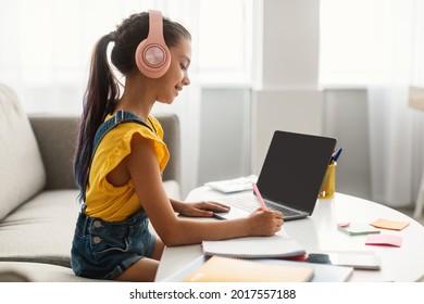 Side view profile portrait of girl in pink headset doing homework, sitting on couch at table using laptop with empty screen, writing essay or making test, taking notes in textbook, free copy space - Powered by Shutterstock