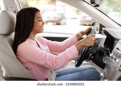 Side view profile portrait of joyful African American woman driving car, doing test drive in dealership showroom. Happy lady riding brand new nice car, sitting on driver's seat in luxury automobile