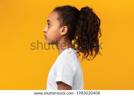 Side view profile portrait of serious black girl in white t-shirt posing isolated on yellow studio background wall and looking at empty free copy space, banner. Person with curly ponytail hair