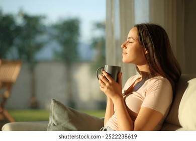 Side view profile of a relaxed woman smelling coffee sitting in a sofa at home at sunset
