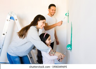 Side View Of A Proud Young Mom And Cute Girl Holding A Paint Roller Together And Painting The Living Room Walls Along With A Smiling Dad