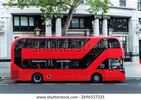 Side view of a red double decker bus stopped on a London street in front of historic stone architecture.