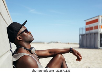 Side View Of Relaxed African Man Leaning To A Wall At Beach Sunbathing. Black Guy Wearing Sunglasses And Cap Relaxing. Afro American Male Model Resting Outdoors