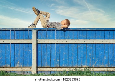 Side View Of Relaxed Boy In Casual Clothes Lying On Blue Hockey Rink Fence And Dreaming Against Cloudy Sky On Summer Day