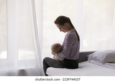 Side View Relaxed Caring Young Mother Breastfeeding Baby Son Daughter Sitting On Bed At Home. Smiling Millennial Woman Delivering Nutrients With Breast Milk To Newborn Child, Enjoying Tender Moment.