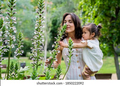 Side View Of Relaxed Mother With Active Child In Hands Smelling Flowers With Closed Eyes And Enjoying Fragrance In Green Park 