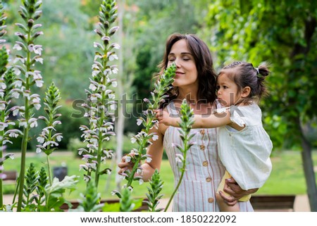 Side view of relaxed mother with active child in hands smelling flowers with closed eyes and enjoying fragrance in green park 
