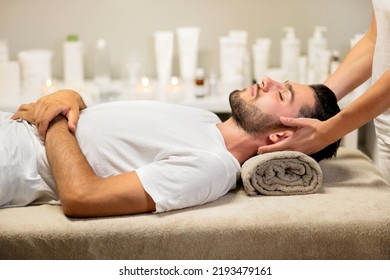 Side view of relaxed young male client in white t shirt closing eyes and lying on rolled up towel while crop masseuse doing neck massage during spa session in beauty salon - Powered by Shutterstock