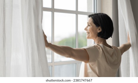 Side view rested middle-aged woman standing near window, open curtains, admires city view, enjoy peaceful, quiet morning, woke up early on weekend, welcoming, start new day, full of hope of happy day - Powered by Shutterstock