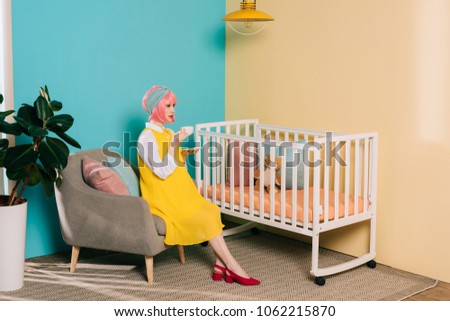side view of retro styled pregnant pin up woman sitting near baby cot with cup of tea in child room