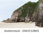 Side view of a rocky mountain on a beach in northern Spain. Blackened, gray and red rock with moss and green grass. Concept of nature