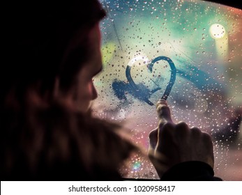 Side View Of Romantic Man Drawing Heart On Steamy Car Window Against Night Lights. 