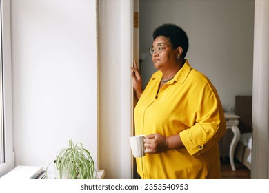Side view of sad african american overweight female in yellow clothes and glasses standing next to window with cup of hot drink, grandma missing her grandchildren, waiting them to visit her - Shutterstock ID 2353540953