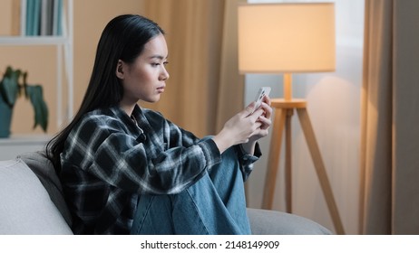 Side View Of Sad Bored Asian Woman Girl Sitting On Sofa At Home With Telephone Serious Unhappy Lady Scrolling Browsing In Smartphone And Using Mobile Phone App Watching Social Media News Feed Typing