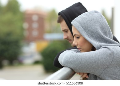 Side View Of A Sad Couple Of Teens Looking Down After Break Up In A Balcony Of A House With An Urban Background