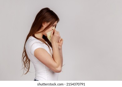 Side view of sad frustrated woman crying and hiding face in hands, upset about loss, bereavement, feeling desperate emotion, wearing white T-shirt. Indoor studio shot isolated on gray background.