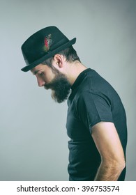 Side view of sad hunched bearded man with hat looking down. Retro toned filtered portrait over gray background with vignette effect. 