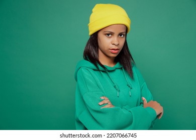 Side View Sad Little Kid Teen Girl Of African American Ethnicity 13-14 Years Old Wear Casual Hoody Hat Hold Hands Crossed Folded Look Camera Isolated On Plain Dark Green Background Childhood Concept