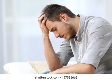 Side View Of A Sad Man With A Hand On The Head Sitting On A Couch In The Living Room At Home