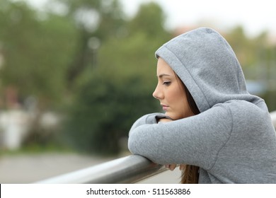 Side view of a sad single teenager looking down in a balcony of her house in a rainy day