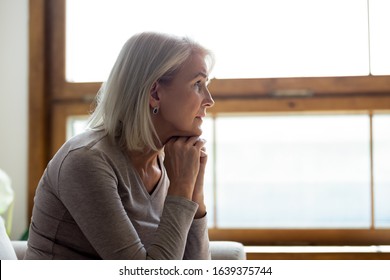 Side View Of Sad Thoughtful Middle-aged Mature Woman Sit On Couch At Home Look In Window Distance Mourning, Upset Pensive Senior Female Lost In Thoughts Thinking Or Pondering Over Past