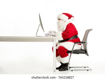 Side View Of Santa Claus Using Computer While Sitting At The Desk Isolated On White