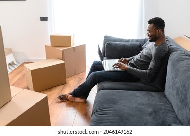 Side View At Satisfied African-American Guy Sitting On The Comfortable Couch And Using Laptop Computer Among Cardboard Boxes, Smiling Black Man Moving Into New Apartment, Shopping Online, Web Surfing