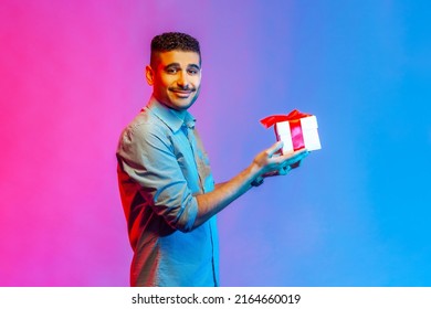 Side View Of Satisfied Delighted Man In Shirt Standing Giving Gift Box, Smiling Looking At Camera Friendly And Generous, Charity. Indoor Studio Shot Isolated On Colorful Neon Light Background.