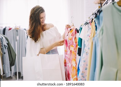 Side view of satisfied female with white paper shopping bags in hands checking price on label on hanger while standing near clothes racks and selecting dress