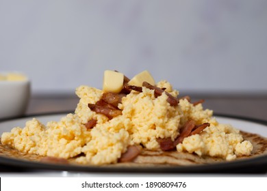 A side view of scrambled eggs and bacon on top of Derbyshire oatcakes sitting on a black rimmed plate. A butter dish is out of focus in the background