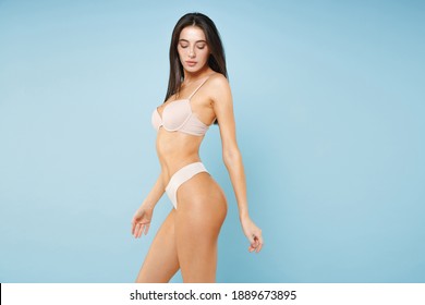 Side view of seductive attractive beautiful young brunette woman 20s in beige underwear showing strong body standing posing looking down isolated on pastel blue colour wall background studio portrait