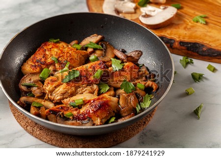 Side view selective focus image of sauteed roasted chicken breasts in a non stick frying pan on white marble seasoned with parsley, spring onion and mushroom as well as herbs, tomato sauce and spices.