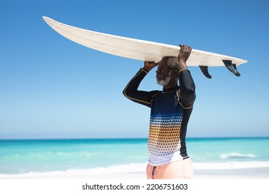 Side view of a senior African American man on a beach in the sun, and holding a surfboard on his head, with blue sky and calm sea in the background - Powered by Shutterstock