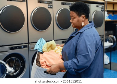 Side view of senior African American woman carrying basket of laundry preparing colored clothes for washing at self service laundromat, copy space - Powered by Shutterstock