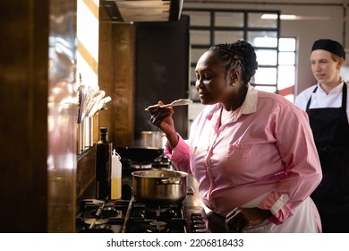 Side view of an Senior African woman at a cookery class, tasting hot vegetables, with a Caucasian woman in the background - Powered by Shutterstock