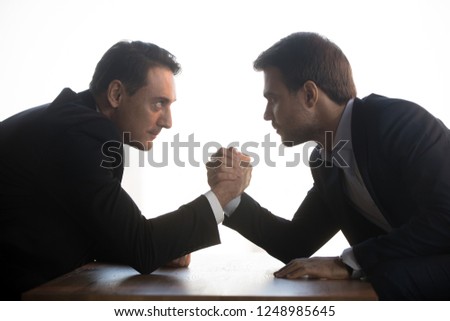 Side view of senior aged and millennial young businessmen in formal suits holds elbows on table arm wrestling at office workplace looking eyes to eyes face to face struggling for leadership at work