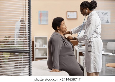 Side view of senior Black woman sitting on therapy bed in medical office breathing out while female health practitioner listening to lung sounds - Powered by Shutterstock