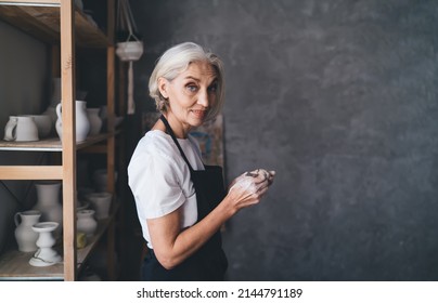 Side view of senior caucasian female sculptor making ceramic pottery with wooden stick and looking at camera. Small business and entrepreneurship. Home hobby, entertainment and leisure. Art studio - Powered by Shutterstock