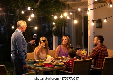 Side View Of A Senior Caucasian Man Standing And Making A Toast Holding A Glass Of Red Wine During A Multi-generation Family Celebration Meal Outside, His Family Is Sitting At The Dinner Table