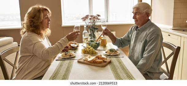 Side View Of Senior European Couple Toasting Wine While Having Dinner At Home. Family Relationship And Spending Time Together. Domestic Lifestyle. Idea Of Romantic Date. Man And Woman Sitting At Table