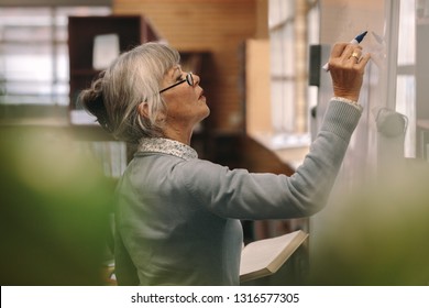 Side View Of A Senior Female Teacher Writing On A White Board In Classroom. Close Up Of A Woman Lecturer Teaching In Classroom.