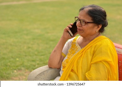 Side View Senior Indian Woman Speaking On Her Smart Phone, Sitting On A Red Bench In A Park In New Delhi. Concept Shot Showcasing Technology Adoption By Senior Citizens. Digital India