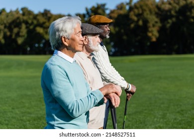 Side View Of Senior Man In Flat Cap Near Interracial Friends With Golf Clubs