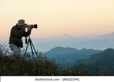 Side view of a senior man taking pictures with camera and telephoto zoom lens on tripod on the mountain peak at sunrise.