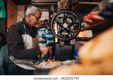 Side view of a senior shoemaker sitting at cobbler's workshop and sewing a leather purse. An old artisan is sitting at sewing machine and using it for repairing a purse. Senior craftsman at his shop. - Powered by Shutterstock