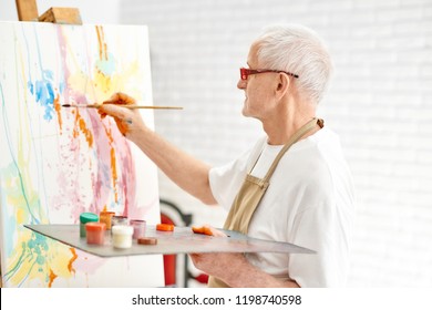 Side view of senior talented painter while painting his masterpiece at bright art studio. Handsome male artist, wearing in white t-shirt and apron and modern glasses drawing abstract picture on easel.