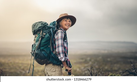 Side View Of A Senior Woman Carrying Backpack Standing On A Hill During Trekking. Smiling Senior Woman Looking Excited During A Hiking Campaign.